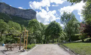 Schotterweg im Campingbereich, links Sitzgruppe unter Pergola, umgeben von Bäumen und Blick auf einen Felsberg.