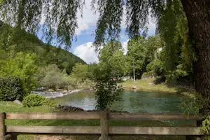 Blick auf einen Fluss mit Holzgeländer, Bäume und Sträucher am Ufer, im Hintergrund grüne Berge und ein Weg.