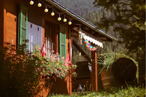 Holzhütte mit Balkon und Blumen vor dem Eingang, im Hintergrund hohe Felswände und Tannen im Alpenpanorama.