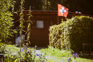Holz-Bungalow auf einem Campingplatz im Grünen, umgeben von Büschen und Blumen, mit Zaun und Flagge am Gartenbereich.