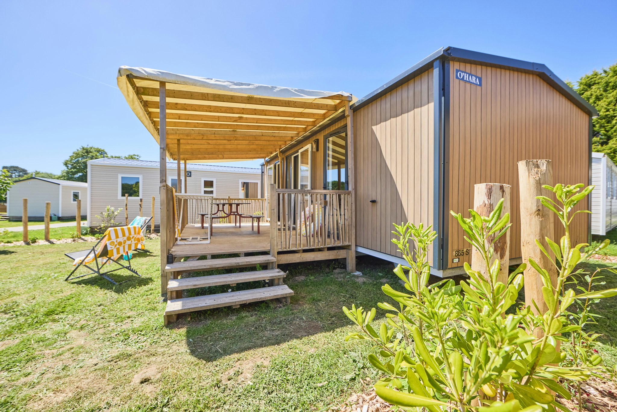 Holzhaus mit Veranda, Stufen und Sitzbank, daneben Sonnenschirm und Liegestuhl auf grünem Rasen.