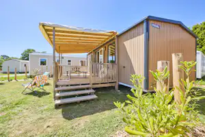 Holzhaus mit Veranda, Stufen und Sitzbank, daneben Sonnenschirm und Liegestuhl auf grünem Rasen.