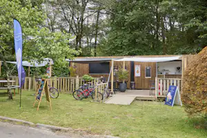 Holzunterkunft mit Terrasse, Fahrrädern am Zaun und Gartenweg, umgeben von Bäumen und grünen Sträuchern.
