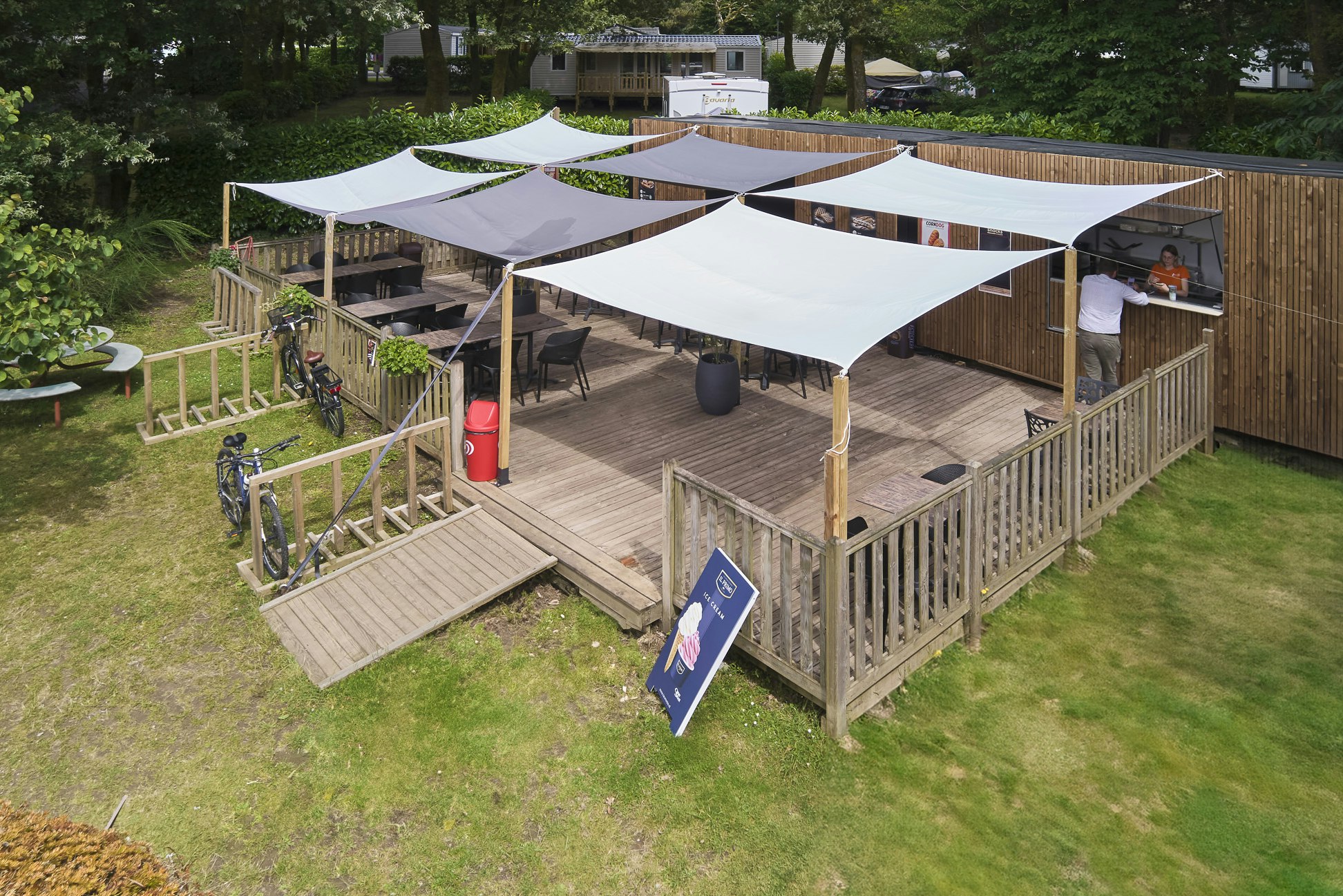 Campingstellplatz mit Holzpodest, mehreren weißen Zeltplanen, Sitzbereich und Fahrrädern im Garten, umgeben von Bäumen und Rasen.
