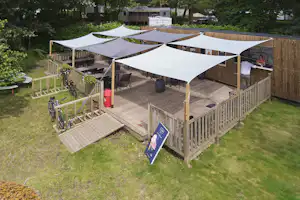 Campingstellplatz mit Holzpodest, mehreren weißen Zeltplanen, Sitzbereich und Fahrrädern im Garten, umgeben von Bäumen und Rasen.