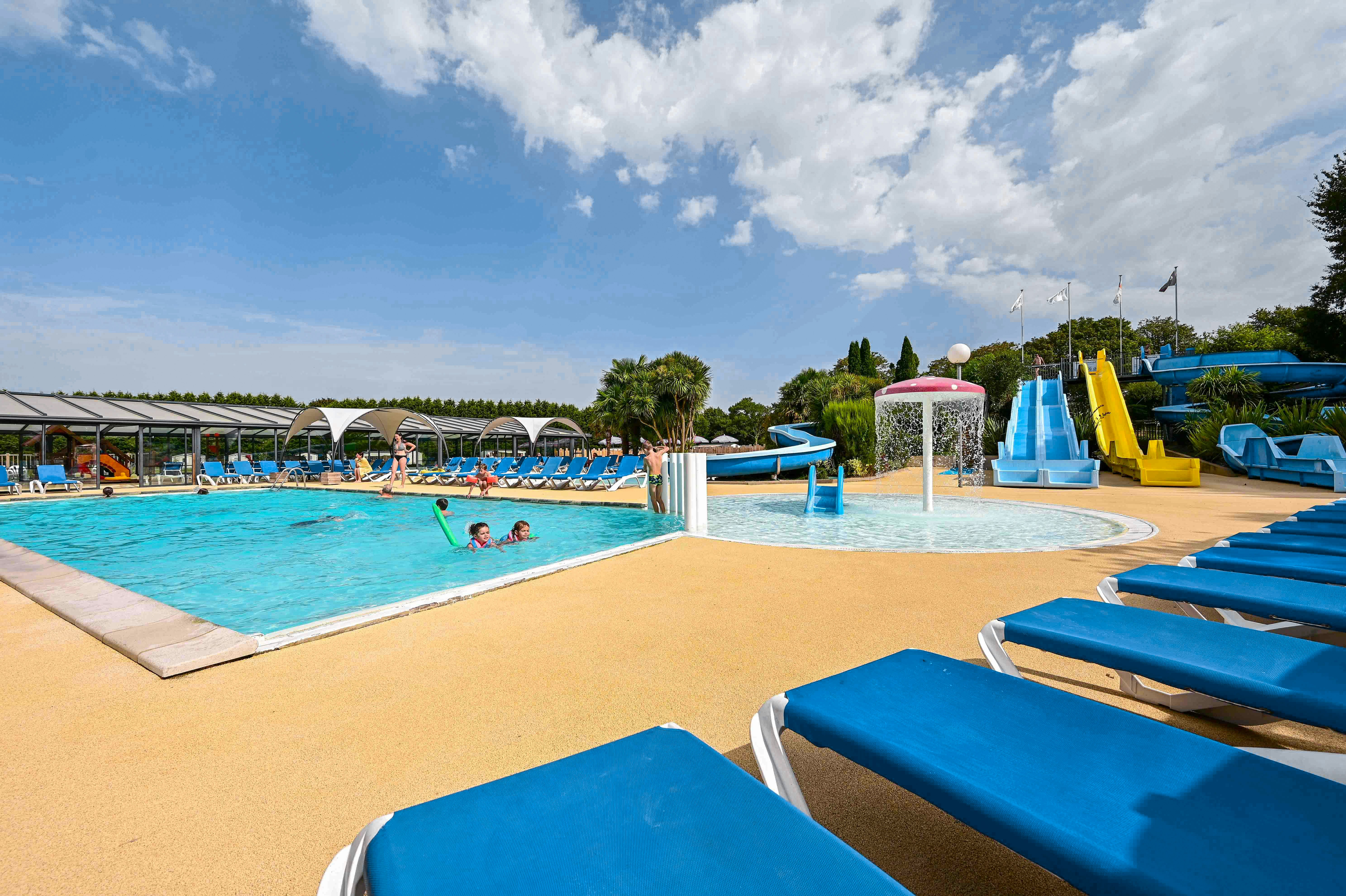 Schwimmbad mit blauen Liegen am Rand, Wasserrutsche und Becken im Hintergrund, Campinganlage mit Sonnenschirmen.