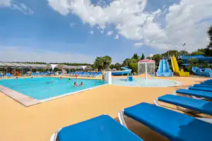 Schwimmbad mit blauen Liegen am Rand, Wasserrutsche und Becken im Hintergrund, Campinganlage mit Sonnenschirmen.