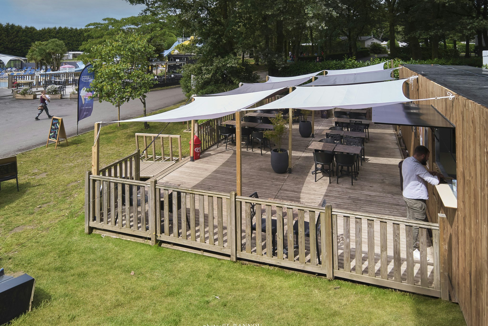 Holzterrasse mit Tisch und Stühlen unter weißen Planen, daneben Rasenfläche und Bäume am Campingplatz.
