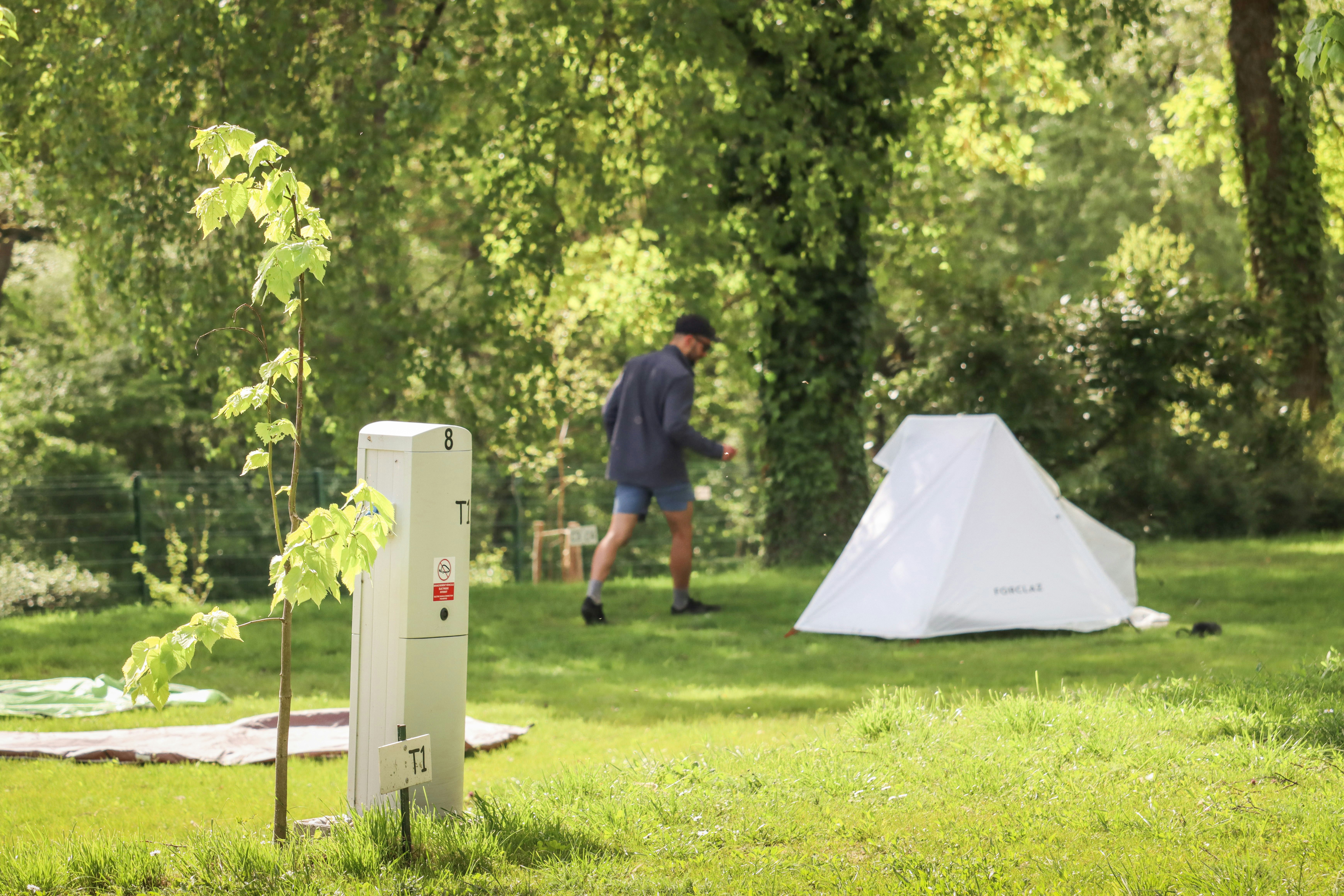 Campingplatz-Parzelle mit Nummer 8, im Hintergrund ein Zelt und ein Mann auf einer grünen Wiese zwischen Bäumen.