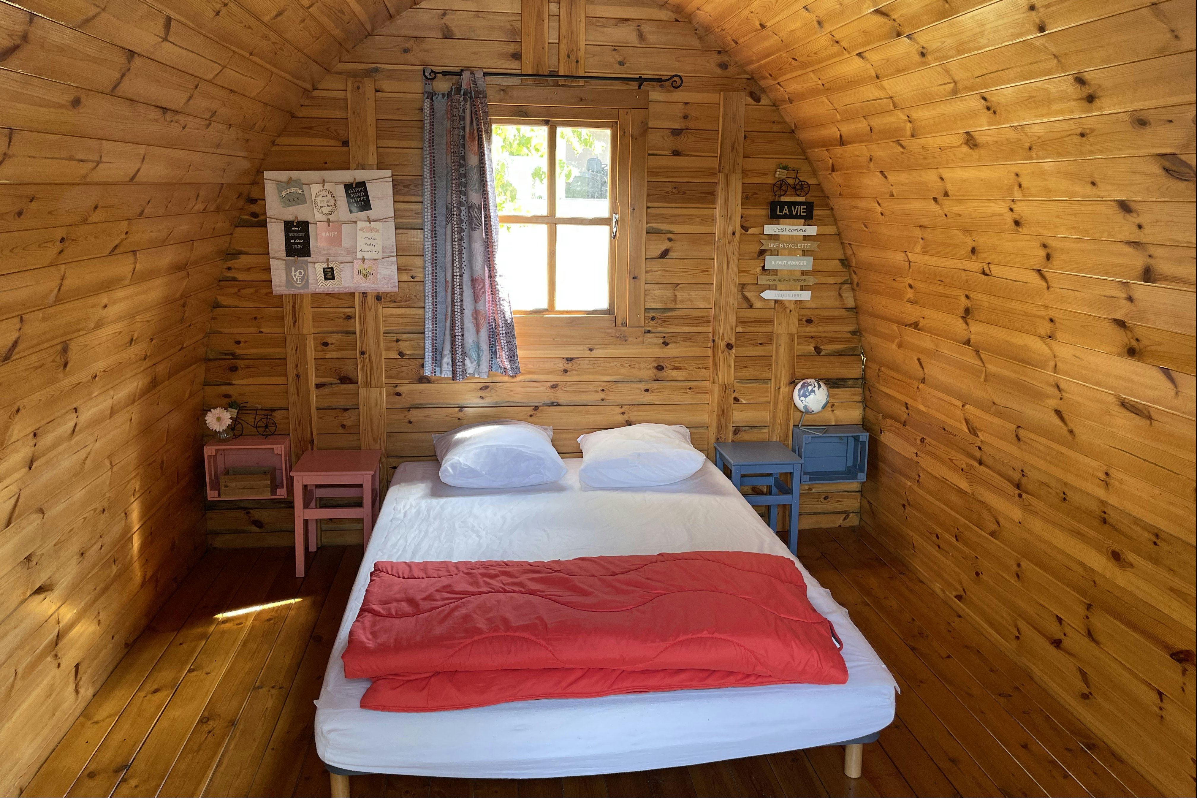 Bett in einer kleinen Holzunterkunft mit zwei Kissen, roter Decke und Fenster im Innenraum, an der Wand Schilder.