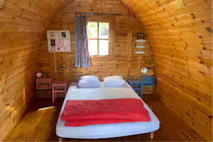 Bett in einer kleinen Holzunterkunft mit zwei Kissen, roter Decke und Fenster im Innenraum, an der Wand Schilder.
