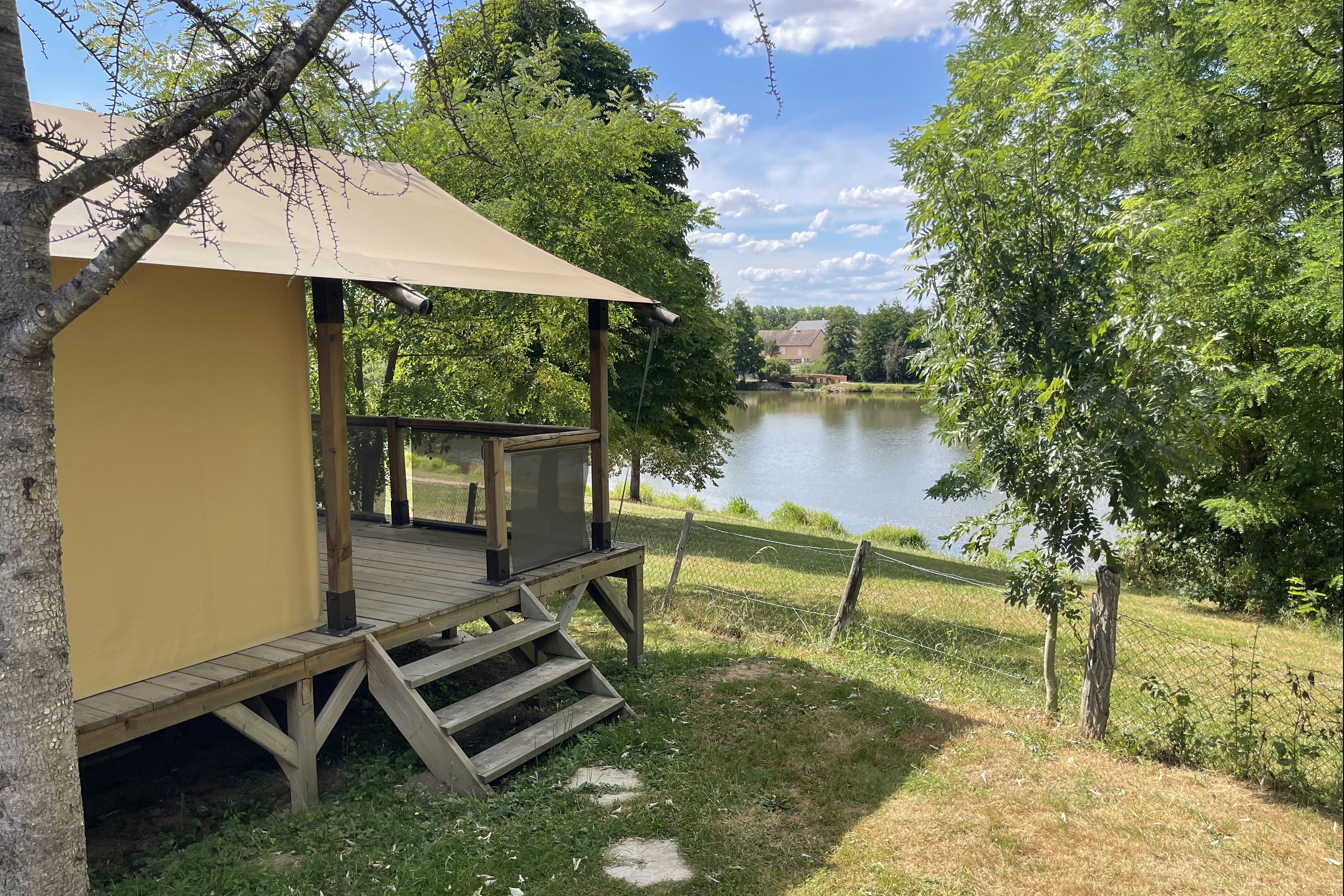 Zelt mit Holzpodest und Veranda am Campingplatz, Blick auf den Fluss durch Bäume und einen gepflegten Rasen.