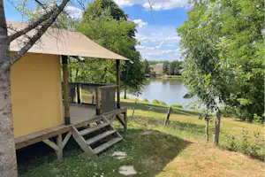Zelt mit Holzpodest und Veranda am Campingplatz, Blick auf den Fluss durch Bäume und einen gepflegten Rasen.