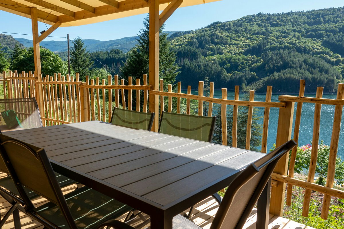 Holzterrasse mit Tisch und Sitzbänken, Blick auf einen See und bewaldete Hügel im Hintergrund.