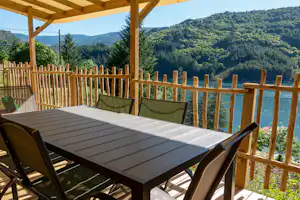 Holzterrasse mit Tisch und Sitzbänken, Blick auf einen See und bewaldete Hügel im Hintergrund.