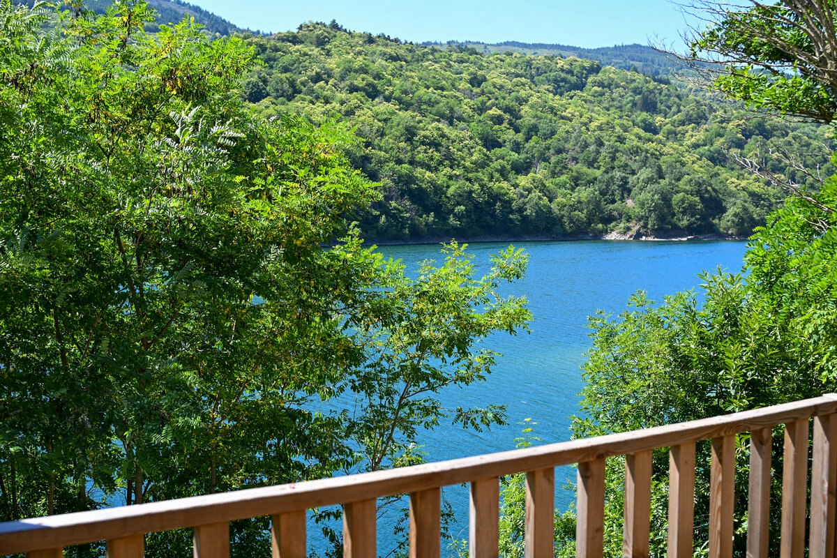 Blick vom Balkon auf einen türkisfarbenen See, umgeben von grünen Bäumen und bewaldeten Hügeln im Hintergrund.