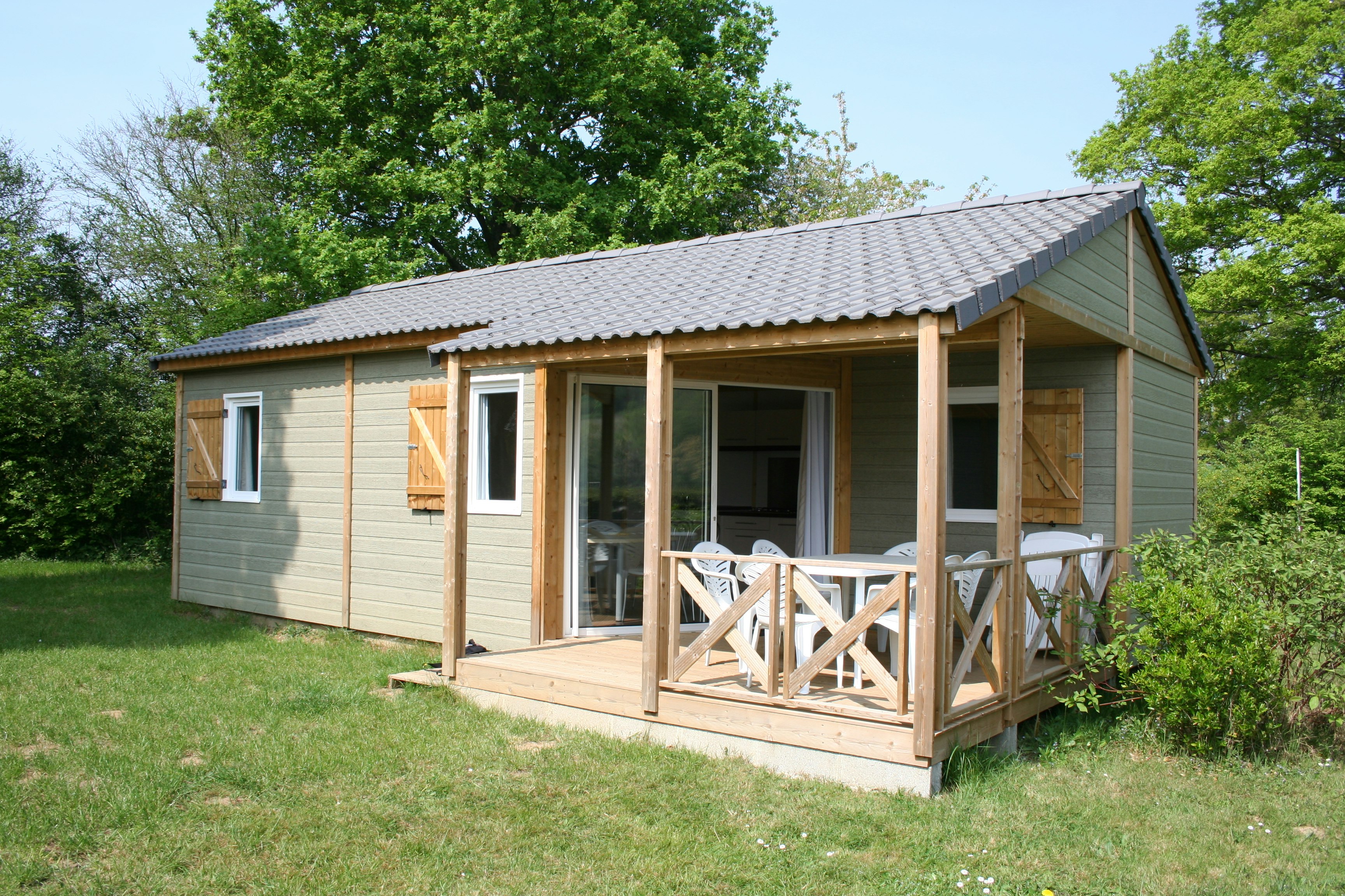 Mobilheim mit Holzterrasse, Sitzgruppe und großen Fenstern, umgeben von Rasen und Bäumen im Grünen.