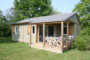 Mobilheim mit Holzterrasse, Sitzgruppe und großen Fenstern, umgeben von Rasen und Bäumen im Grünen.