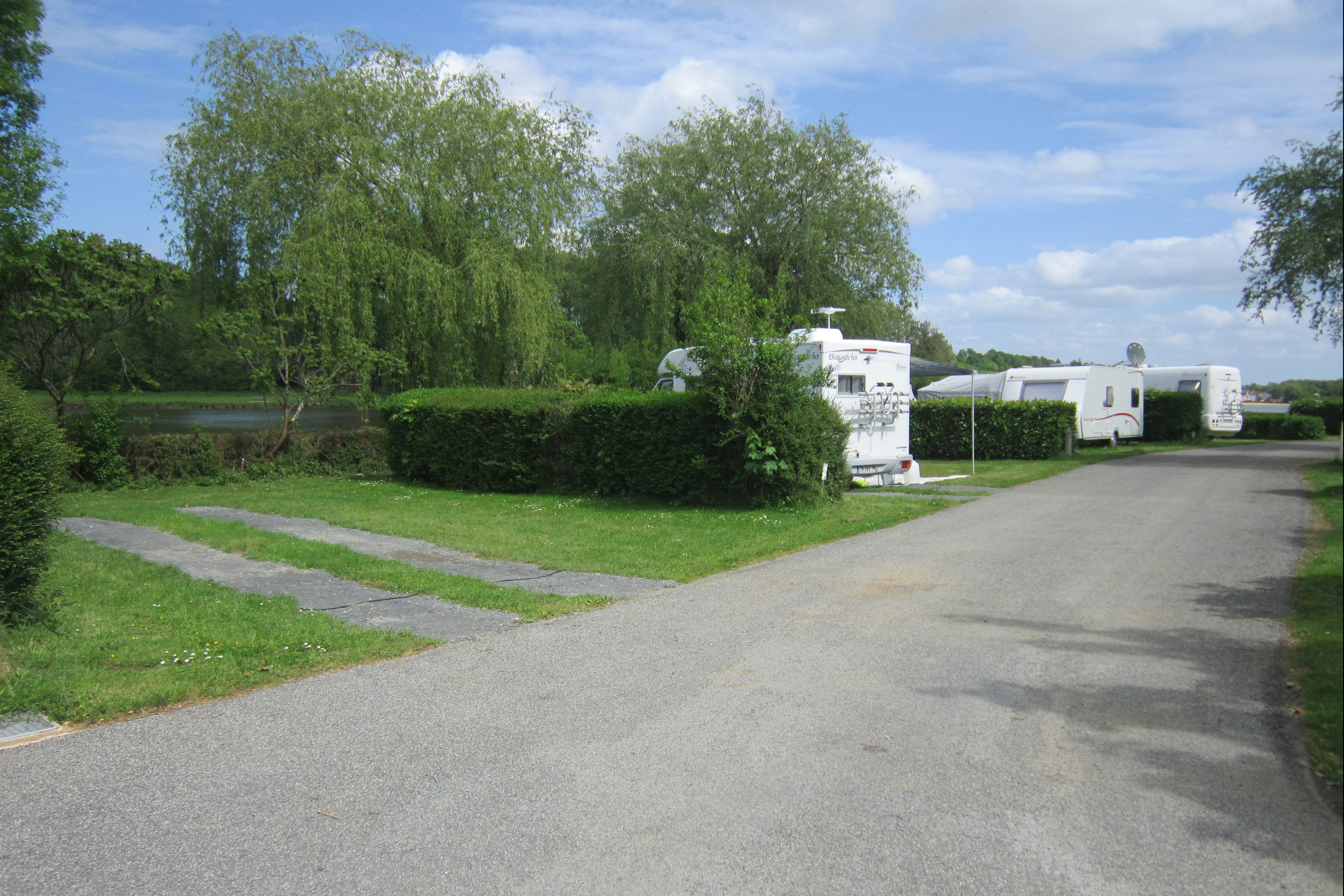 Wohnwagen auf einem Campingplatz-Fahrweg, Hecken und Bäume am Rand, asphaltierter Weg und Stellplätze in der Ferne.