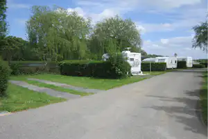 Wohnwagen auf einem Campingplatz-Fahrweg, Hecken und Bäume am Rand, asphaltierter Weg und Stellplätze in der Ferne.