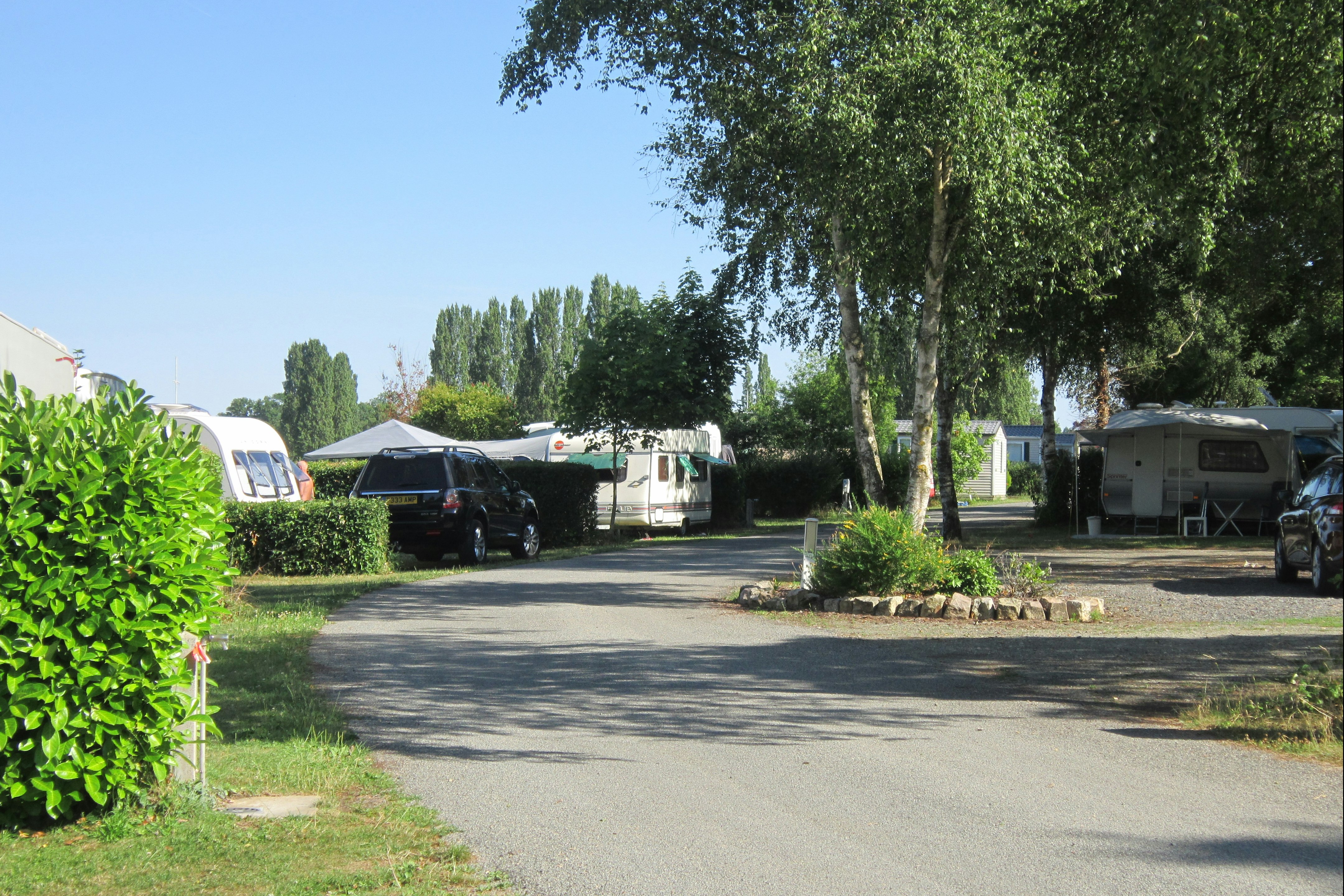 Campingplatz-Allee mit Wohnwagen und Autos, Bäume und Hecken am Wegesrand, Stellplätze entlang der Straße.