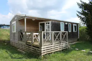 Holzferienhaus mit Veranda, großen Fenstern und Holzgeländer auf einem Campingplatz, umgeben von Wiese und Bäumen.