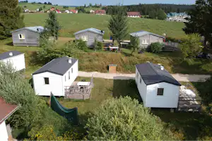 Mehrere Ferienhäuser auf einem Campingplatz mit Holzterrassen, Tischen und Stühlen, umgeben von Bäumen und grünen Wiesen.