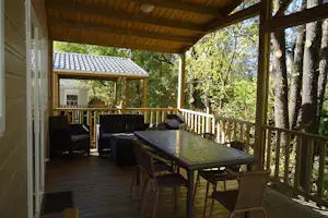 Holzterrasse mit Esstisch und Gartenstühlen unter einer Überdachung, im Hintergrund ein kleines Gebäude und Bäume.
