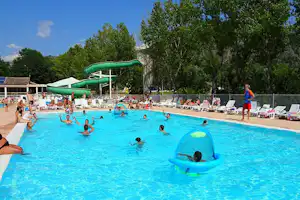 Großes Camping-Schwimmbad mit blauer Wasserfläche, grüner Rutsche, aufblasbarem Spielzeug und Liegestühlen unter Bäumen.