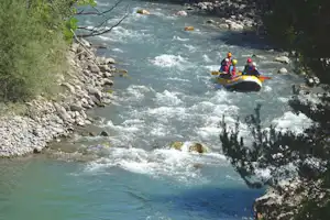 Rafting-Boot mit mehreren Personen auf einem reißenden Fluss, umgeben von Felsen und Bäumen am Ufer.