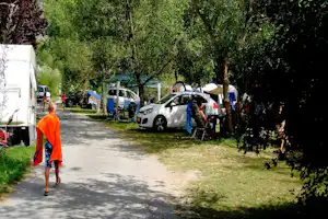 Camping-Gasse mit Wohnwagen und Autos entlang des Wegs, Bäume spenden Schatten auf dem Stellplatz.