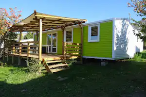 Grüner Wohnwagen mit Holzveranda und Treppe auf dem Campingplatz, umgeben von Rasen und Bäumen.