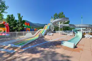 Wasserrutsche und Pool auf einer Camping-Terrasse mit Liegen, bunten Spielgeräten und Blick auf die Berge.