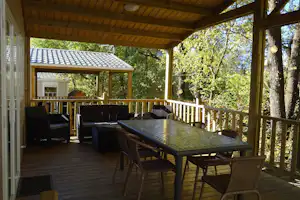 Überdachte Camping-Terrasse mit Tisch und Stühlen, Blick in den Garten mit Bäumen und Holzgeländer.