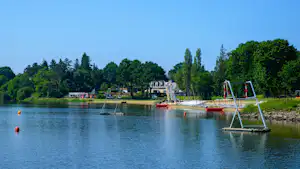 Blick auf den See mit Campingplatz am Ufer, Schaukeln und roten Booten, im Hintergrund Bäume und Gebäude.