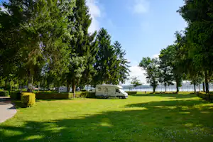 Wohnmobil auf einem Campingplatz zwischen hohen Bäumen, mit Rasenfläche und Hecke am Weg.