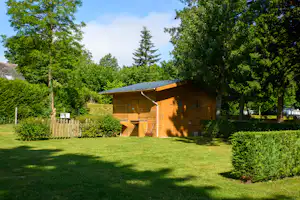 Holz-Blockhaus auf einem Campingplatz mit Rasen, Hecke und Bäumen im Hintergrund.