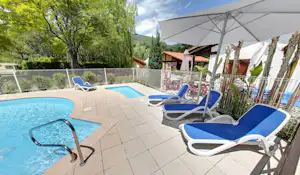 Camping-Pool mit blauem Wasser, mehreren Liegen und Sonnenschirm, daneben Kacheln und Zaun, im Hintergrund Gebäude und Bäume.