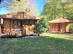 Zwei Holzbungalows auf einem Campingplatz mit Terrasse, Holzgeländer und Rasen, umgeben von Bäumen.