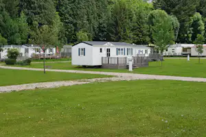 Wohnwagen mit Holzterrasse und weißen Fenstern auf einem Campingplatz, umgeben von Rasen, Wegen und Bäumen im Hintergrund.