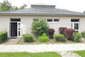 Campinggebäude mit Eingangstür, Fenstern und gepflegten Heckenbeeten vor dem Haus, gepflasterter Weg und Rasenfläche.