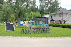 Reihe Fahrräder am Campingplatz neben Spielgeräten und Trampolin, im Hintergrund Wohnhaus und viel Grün.