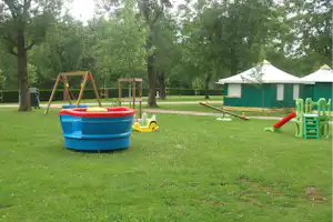 Spielplatz auf dem Campingplatz: Planschbecken, Schaukelgestell und Rutsche neben grünen Zelten im Schatten von Bäumen.