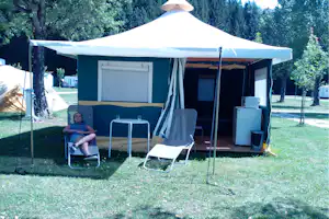 Camping-Unterkunft mit Zeltveranda, Sitzgruppe und Tisch vor dem Eingang, umgeben von Bäumen und Rasen.