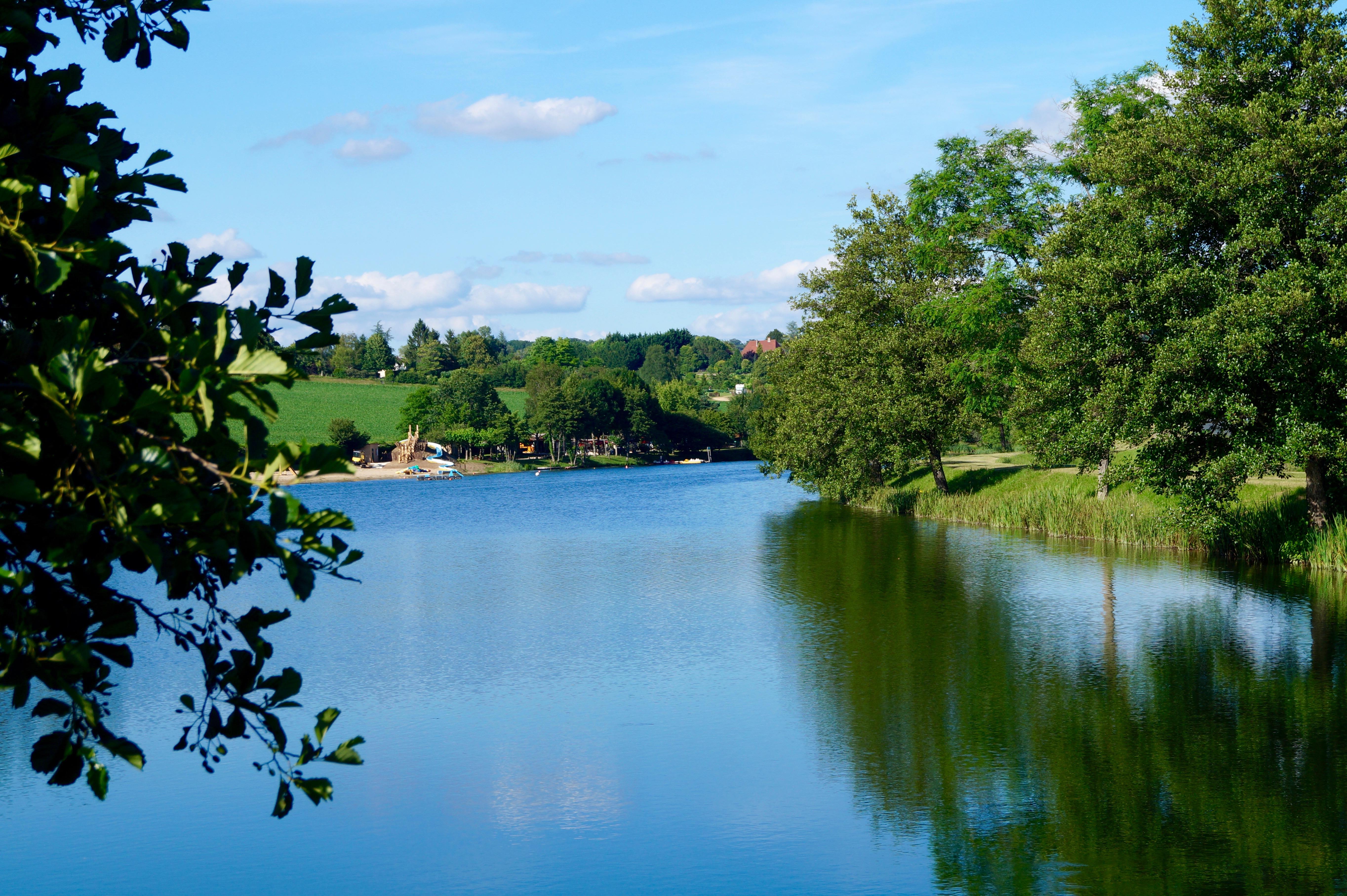 Ruhiger See mit spiegelnder Wasseroberfläche, grünen Bäumen und Uferweg, Blick vom Campingplatz Richtung Wald und Häuser.