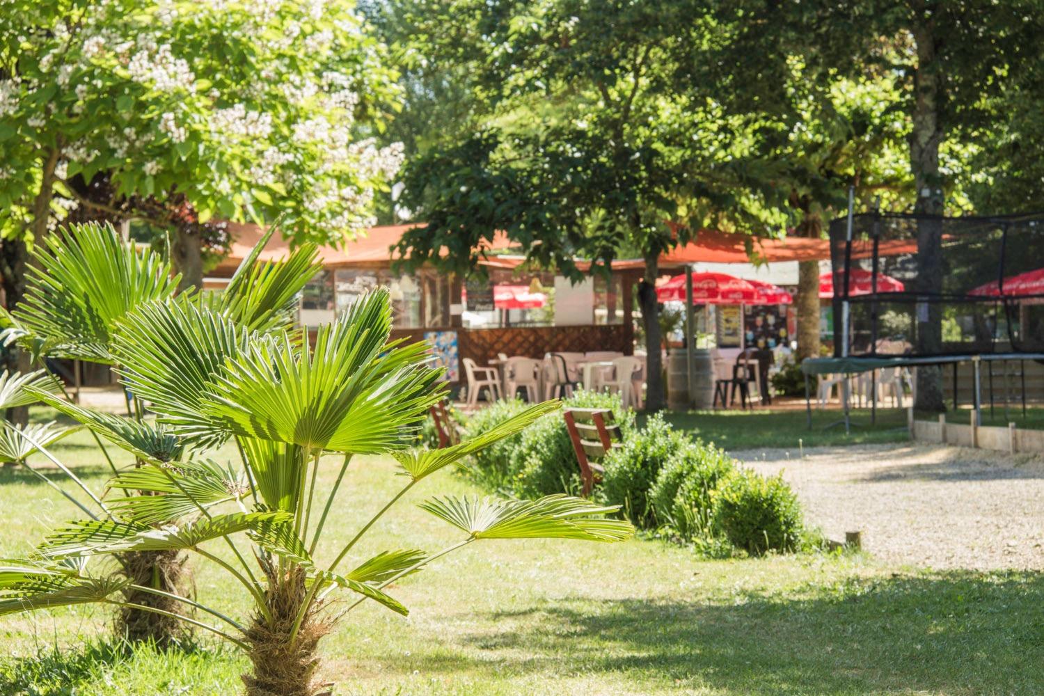 Garten mit Palmen, Kiesweg und Sitzplätzen auf einer Terrasse vor einem Restaurant am Campingplatz.