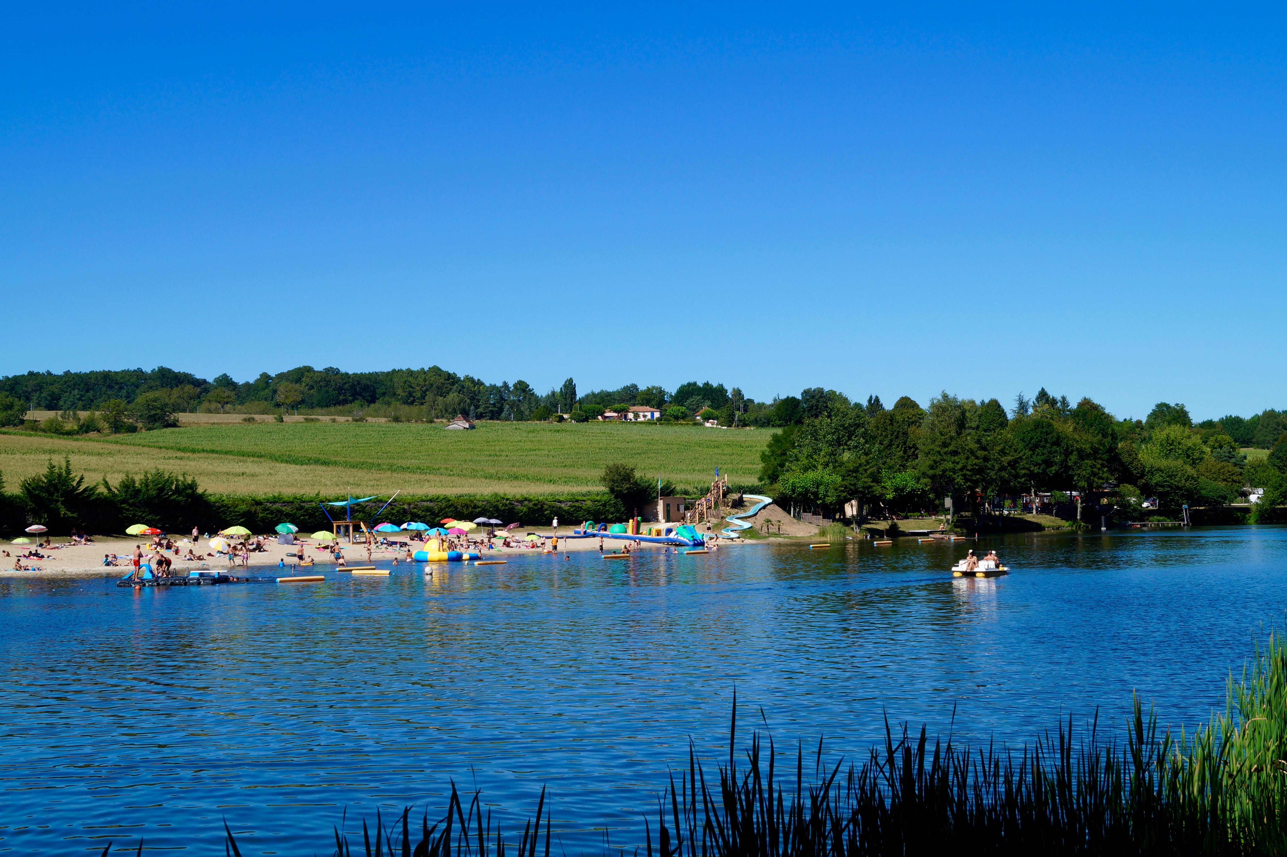 Blick auf einen See mit Campingplatz am Ufer, Booten im Wasser und Liegewiese, im Hintergrund grüne Landschaft und Häuser.