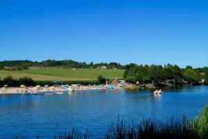 Blick auf einen See mit Campingplatz am Ufer, Booten im Wasser und Liegewiese, im Hintergrund grüne Landschaft und Häuser.
