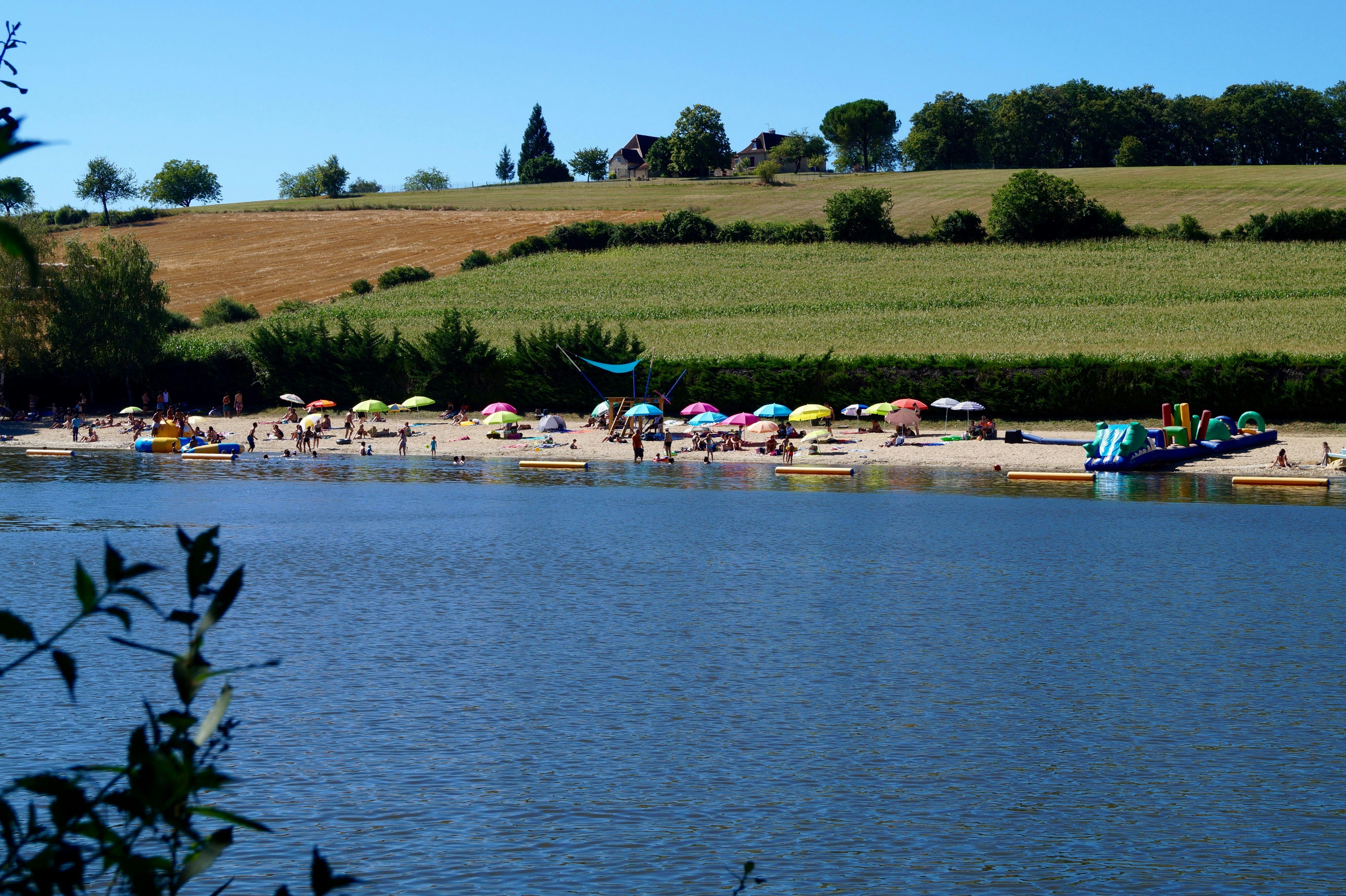 Blick auf einen See mit Liegen und Sonnenschirmen am Ufer, im Hintergrund Wiesen und Bäume.