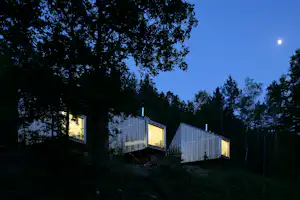 Campingzelte mit transparenten Wänden zwischen Bäumen am Abend, im Hintergrund ein beleuchtetes Gebäude und der Himmel.